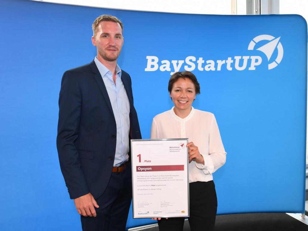 Zwei Personen Gewinnerteam Opsyon vor dem BayStartUP - Logo