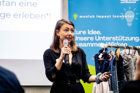 Munich Impact Incubator 2024 - Gründerin spricht, hinter ihr ein Ständer mit Kleidung