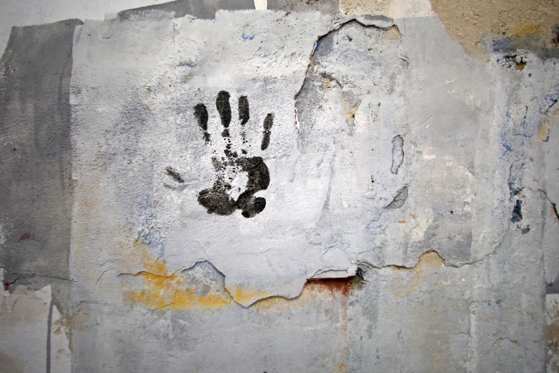Eine rauhe, alt anmutende Wand, die mit hellen Farben künstlerisch bemalt ist und mit dem schwarzen Farbabdruck einer Hand verziert ist.
