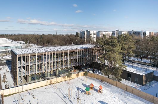 Die bereits im Bestand vorhandenen Kiefern konnten trotz der Nähe zum Neubau erhalten werden. Im Frühling muss der Rasen in der Freispielfläche anwachsen. Bis dahin bleibt der Bauzaun noch stehen. Danach – wie auf dem Bild gut zu erkennen ist – wird den Kindern eine richtig große Fläche mit Spielgeräten, Wasserspiel und Sandkästen zur Verfügung stehen.