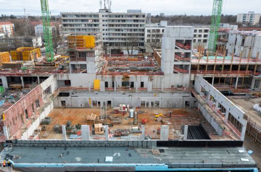 Ansicht auf die Grundschule mit Holzstützen, das Verbindungsgebäude mit Mensa und Pausenhalle, das Erdgeschoss der Mittelschule und im Vordergrund auf die Sporthalle (von rechts nach links).