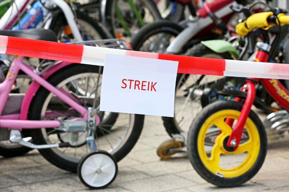 Das Bild zeigt im Hintergrund mehrere Kinderfahrräder. Vor den Fahrrädern befindet sich ein rot-weißes Absperrband. Daran geheftet ist ein A4-Blatt mit der Aufschrift 