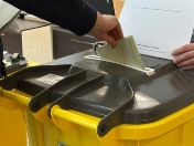 Eine Person wirft einen Stimmzettel in eine Wahlurne, während eine zweite Person daneben steht und Unterlagen hält.