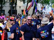 Teilnehmende des Christopher Street Day in München stehen in einer Menschenmenge und tragen dunkelblaue T-Shirts der Feuerwehr München mit Regenbogenmotiv. Im Hintergrund sind zahlreiche Pride-Flaggen und Zuschauer*innen entlang der Straße zu sehen.
