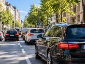 Schwarzer Pkw fährt auf einer mehrspurigen, von Bäumen gesäumten Straße im Stadtverkehr. Weitere Fahrzeuge sind zu sehen.
