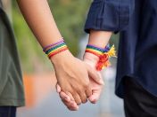 Zwei Personen halten Händchen. Beide tragen Armbänder in Regenbogenfarben – ein Symbol für LGBTIQ*-Stolz und Vielfalt.