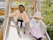 Zwei lachende Kinder rutschen nebeneinander eine breite Rutsche auf einem Spielplatz hinunter.