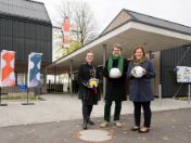 Bild: Vor der neuen Sportanlage an der Grohmannstraße stehen drei Personen mit Bällen in den Händen: Baureferentin Jeanne-Marie Ehbauer, Stadtschulrat Florian Kraus und Bürgermeisterin Verena Dietl. Im Hintergrund sind Fahnen der Landeshauptstadt München zu sehen