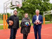 Bild:  Sportreferent Florian Kraus, Jeanne-Marie Ehbauer und Oberbürgermeister Dieter Reiter (v.l.n.r.) stehen auf einem neu gestalteten Außenspielfeld mit Basketballkörben. Jeder von ihnen hält einen Ball in der Hand – Basketball, Volleyball und Fußball – und lächelt in die Kamera. Im Hintergrund sind grüne Bäume, Rasenflächen und weitere Sporteinrichtungen zu sehen.