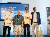 Teilnehmende der campus42-Eröffnung (Mathias Marschall (Leitung PIZKB), Florian Kraus (Stadtschulrat), Sebastian Weisenburger (Stadtrat), Dr. Sonja Moser (Leitung campus42)) posieren mit Stofftaschen vor einer Präsentation.
