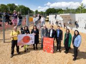 Bild: Mehrere Erwachsene stehen bei der Auftaktfeier zur Schulhoföffnung auf einem Schulsportplatz. Im Hintergrund spielen Kinder an einer Kletterwand.
