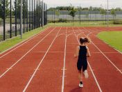 Eine Läuferin in schwarzer Sportkleidung läuft auf einer roten Tartanbahn im Sportpark. Im Hintergrund sind weitere Sportflächen, Zäune, Bäume und ein heller Himmel zu sehen.
