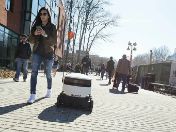 Frau läuft neben mobilem Roboter zwischen Menschen die Straße entlang