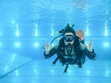 Bild: Eine Person taucht in einem Schwimmbecken mit kompletter Tauchausrüstung, inklusive Maske, Atemgerät und Sauerstofftank. Sie zeigt mit beiden Händen das „OK“-Zeichen unter Wasser.