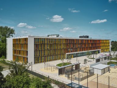 Auf dem Bild ist die Grund- und Mittelschule Rockefellerstraße aus der Vogelperspektive zu sehen. Vor der Schule ist der Pausenhof und Spiellplatz zu sehen. 