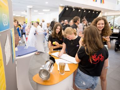 Eintrag in das Gästebuch der Einweihung neben der Zeitkapsel zum Befüllen mit Wünschen für die neue Schule. 4 Schülerinnen stehen an einem Infostand neben der Bühne und eine trägt sich in das Gästebuch ein.