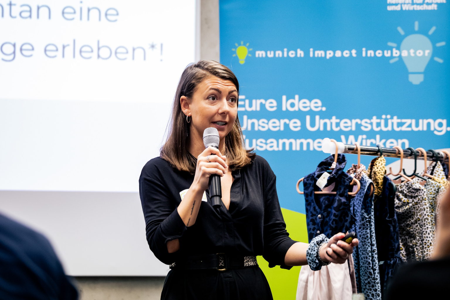 Munich Impact Incubator 2024 - Gründerin spricht, hinter ihr ein Ständer mit Kleidung